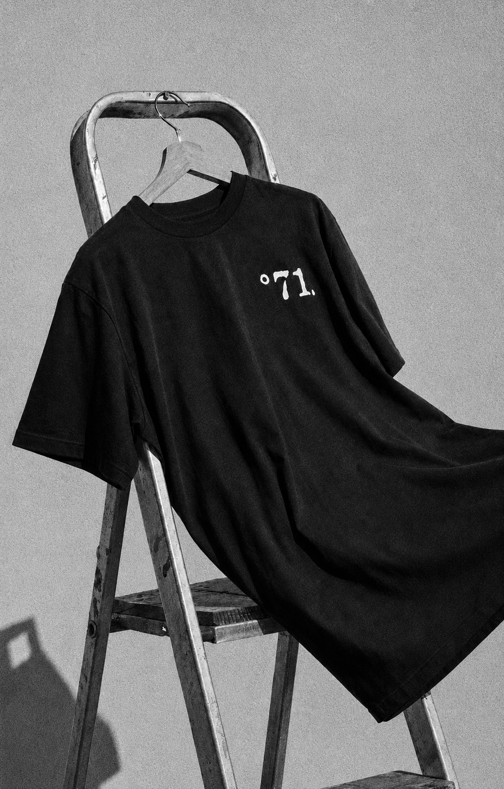 A black oversized short-sleeve T-shirt with a small white ”°71.” chest graphic, draped over a metal stepladder against a gray wall.