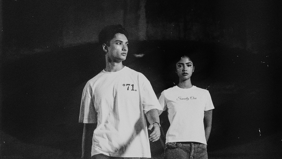 Black and white photo of two models wearing white graphic t-shirts, standing in a dark, moody setting. The shirt on the left features the text '°71.