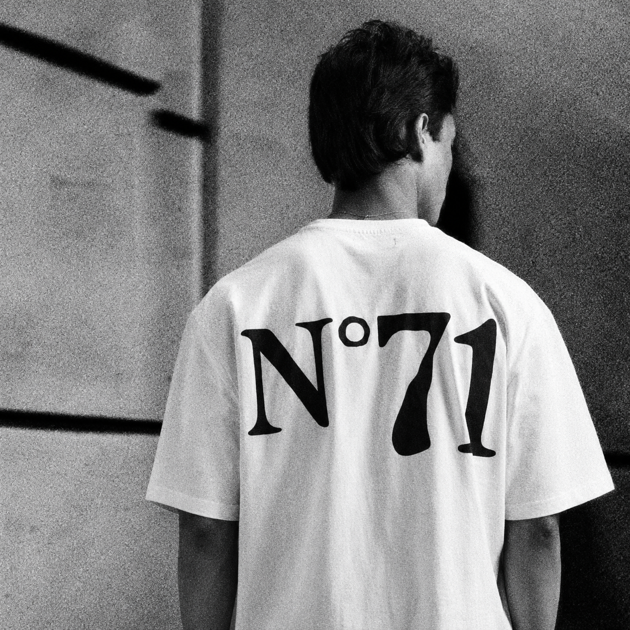 A black and white photo showing the back of a person wearing a white t-shirt with a large black 'N°71' graphic printed on the back, standing against a textured wall.