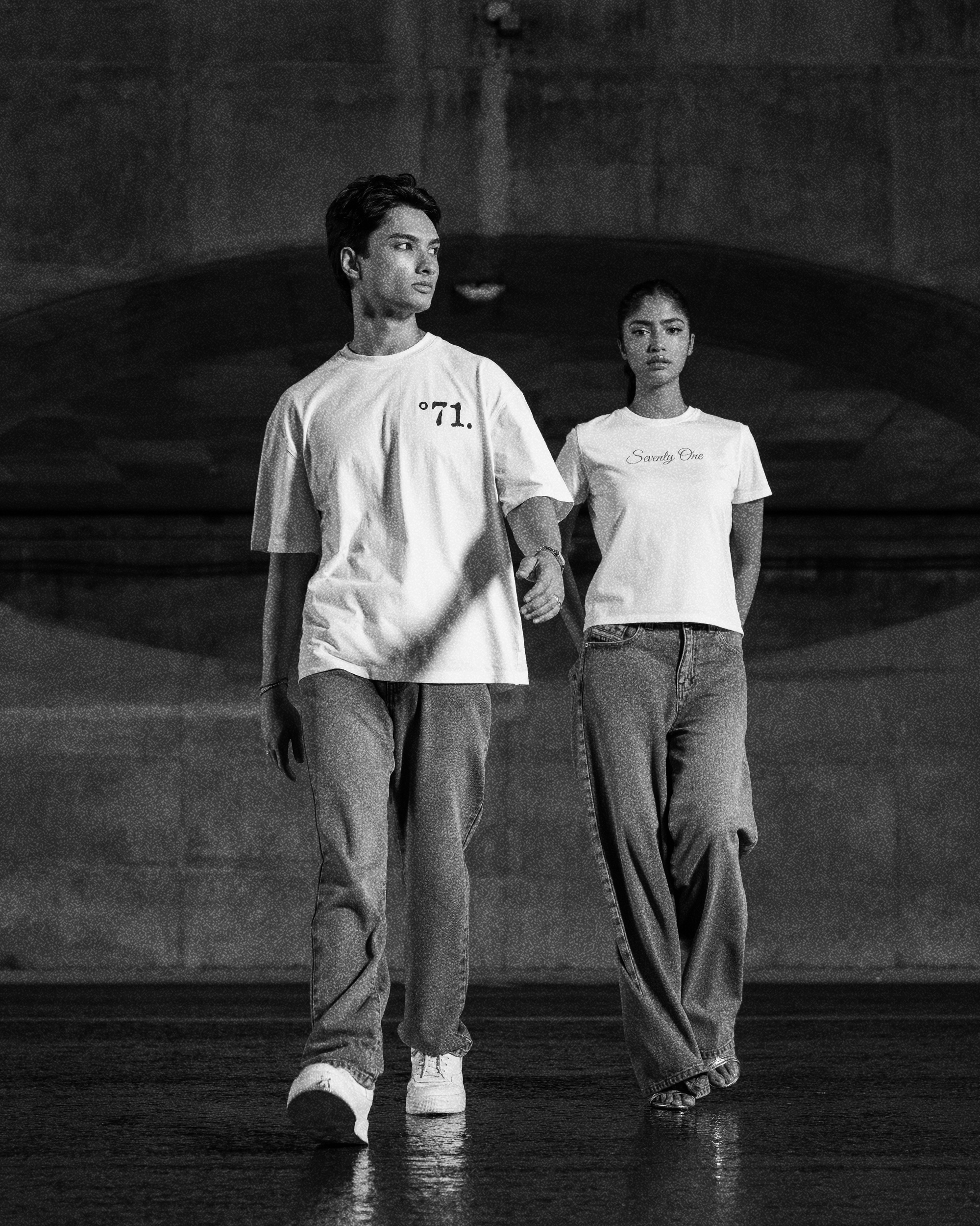A black and white fashion photograph of a young man and woman walking arm-in-arm in front of a concrete archway. The man wears an oversized white t-shirt with '71.