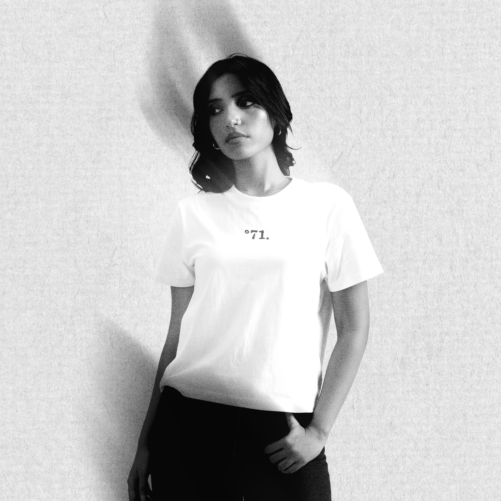 A woman wearing a white short-sleeve t-shirt with a small black typewriter-style graphic reading '°71.' on the chest, paired with black pants, standing against a textured light gray wall.