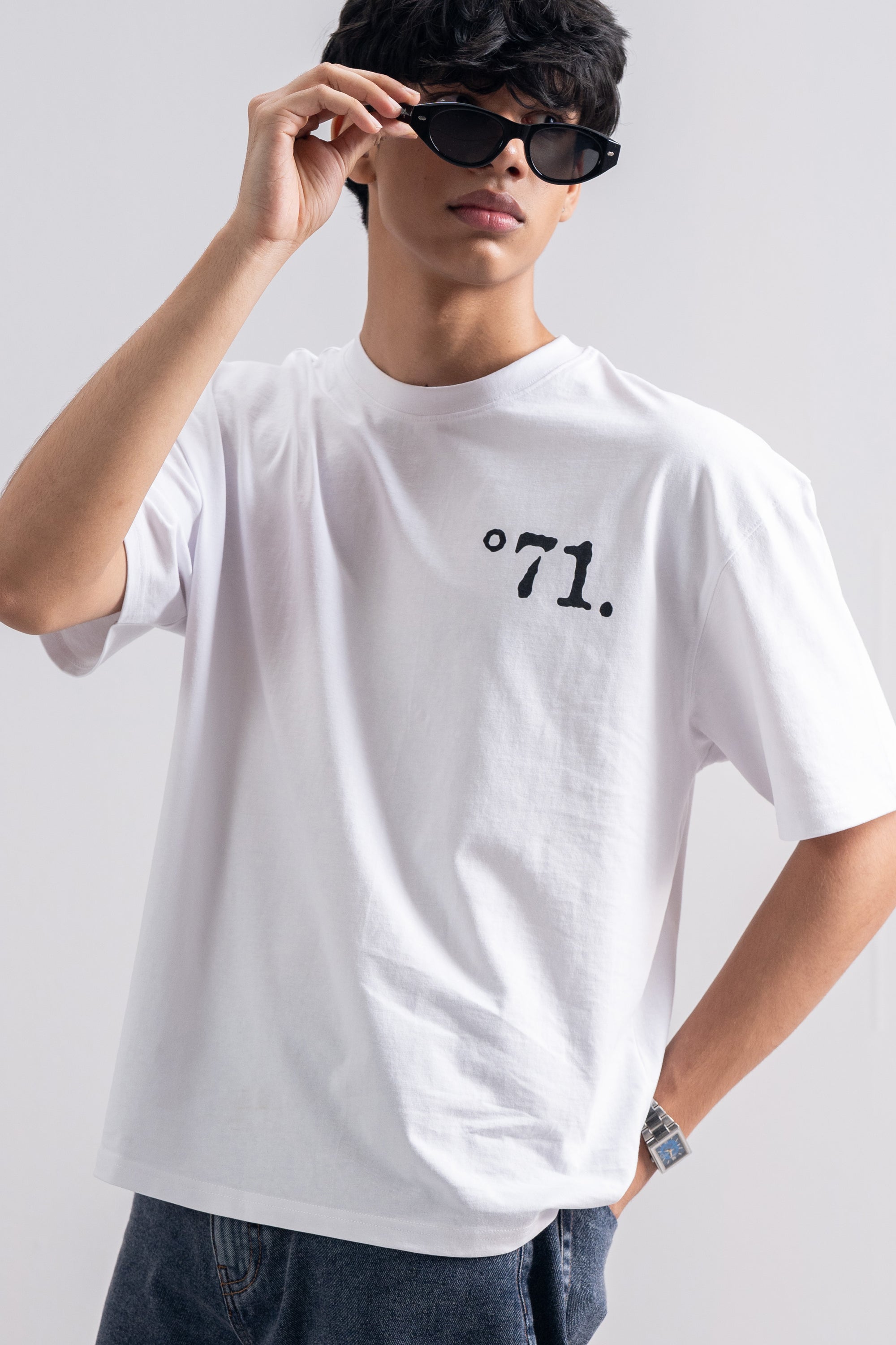 A man wearing a white crewneck t-shirt with a black printed '°71.' logo on the chest, styled with dark sunglasses and blue jeans against a light gray background.