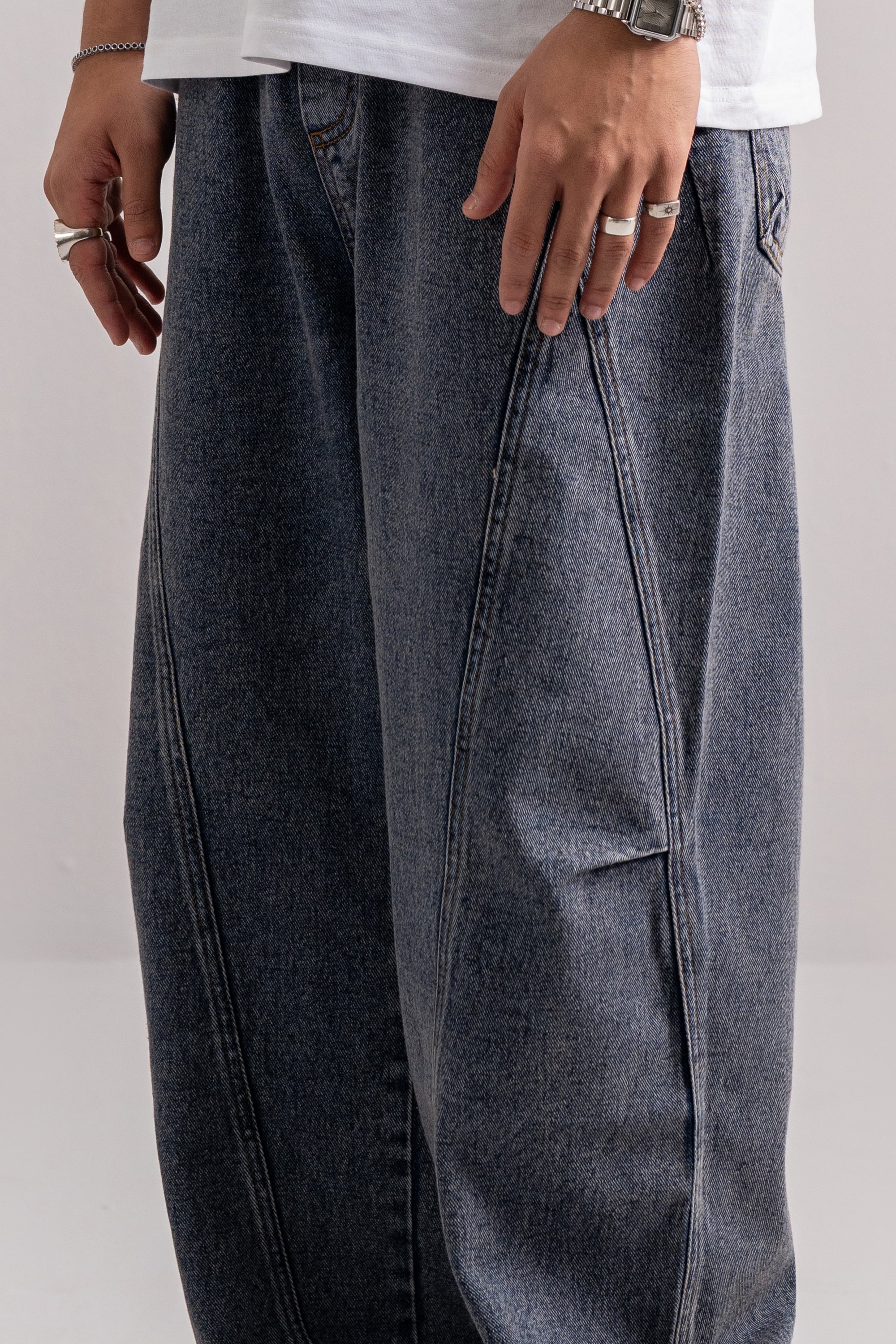 Close-up of a person wearing very wide-leg dark gray denim jeans with diagonal seam details, paired with a white t-shirt. The person is wearing silver rings and a watch.