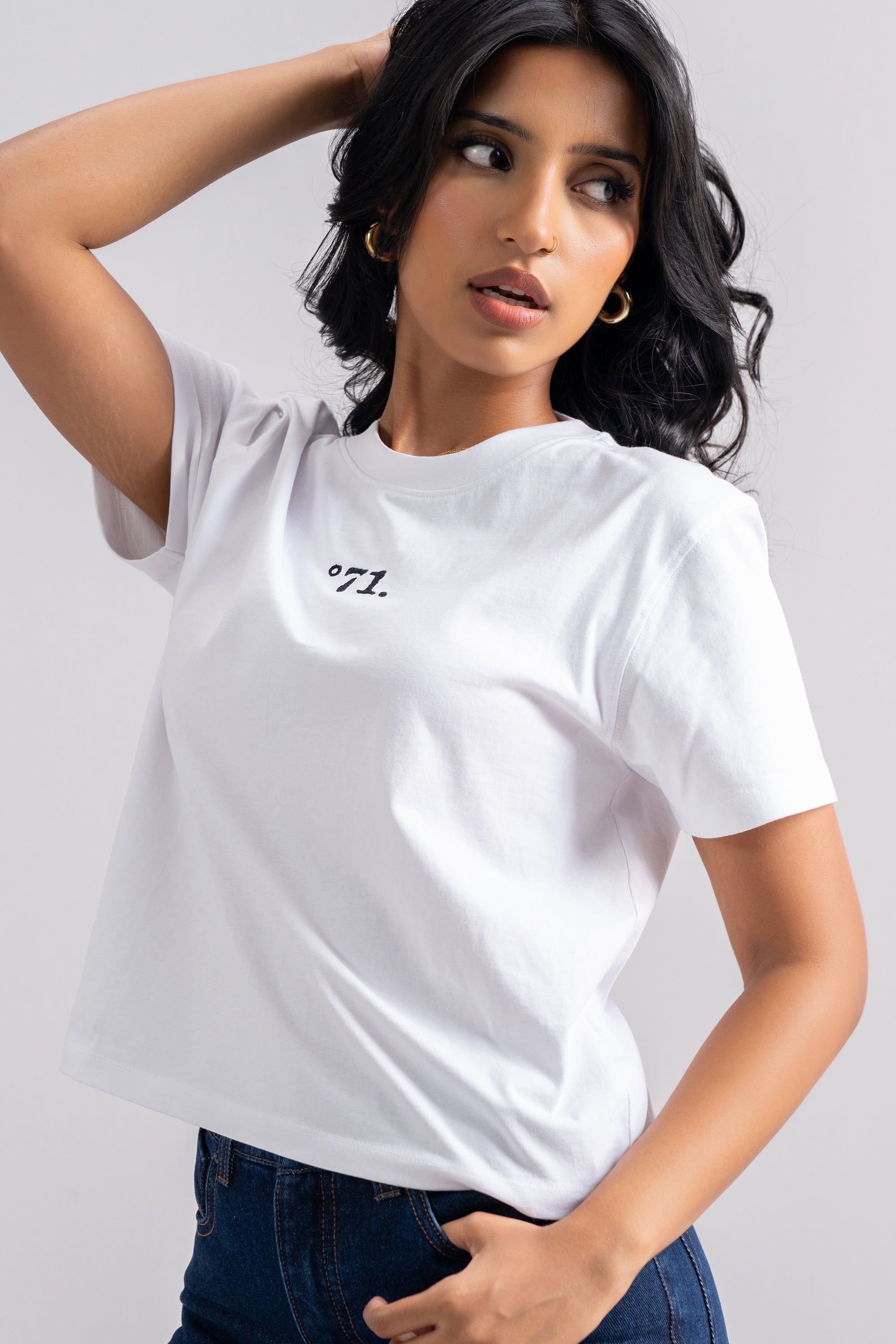 A woman wearing a white short-sleeve crop top t-shirt with a small black printed logo on the left chest, paired with dark blue jeans, against a light gray studio background.