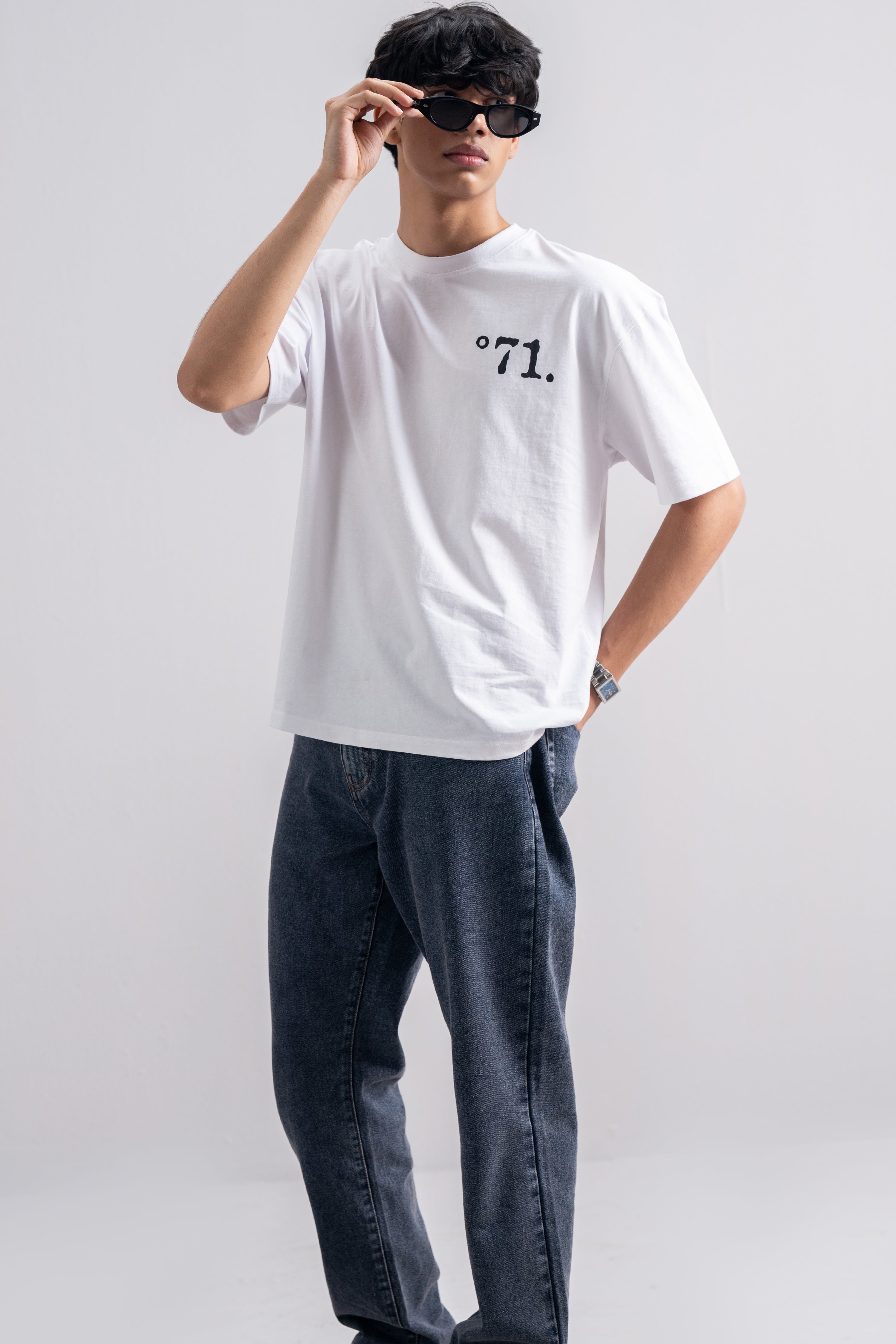 A man modeling a white short-sleeve crewneck t-shirt with a black printed logo reading '°71.' on the left chest, paired with dark blue jeans and black sunglasses, against a light gray studio background.