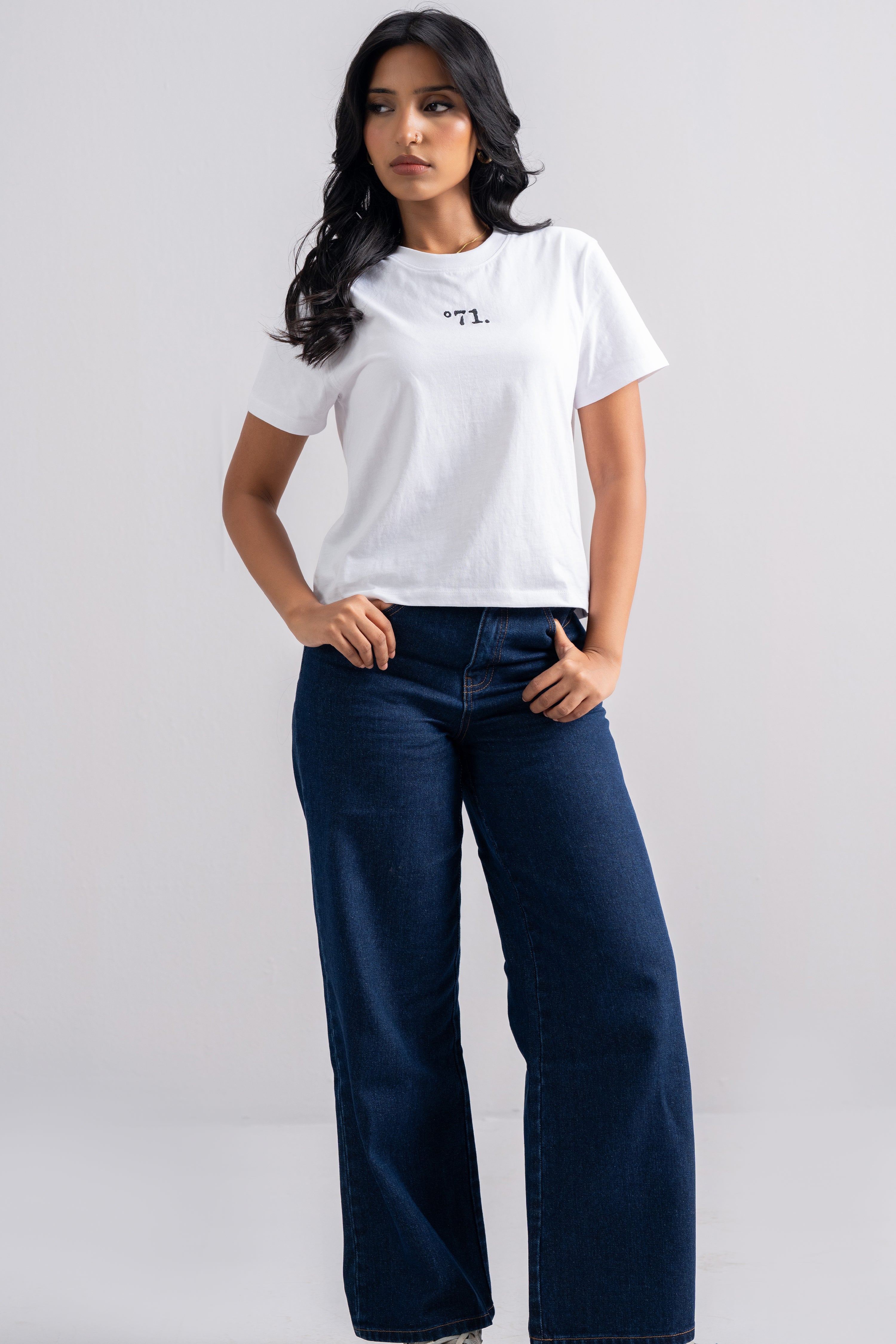 A woman modeling a white short-sleeve t-shirt with a small black graphic on the chest, paired with dark blue wide-leg jeans, against a light gray studio background.