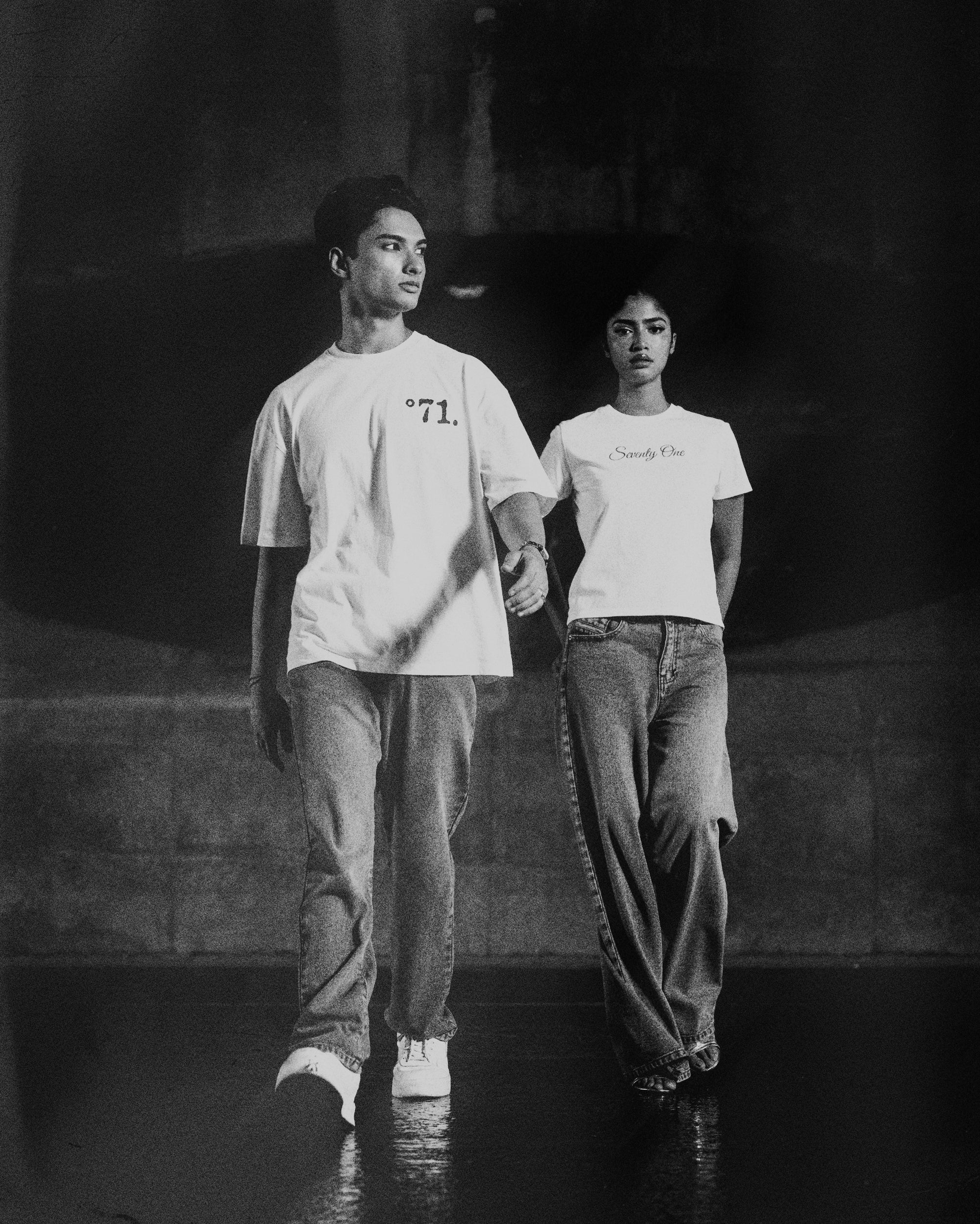 A black and white fashion photograph of two models walking hand-in-hand on a dark runway. Both are wearing oversized white graphic t-shirts and wide-leg jeans.