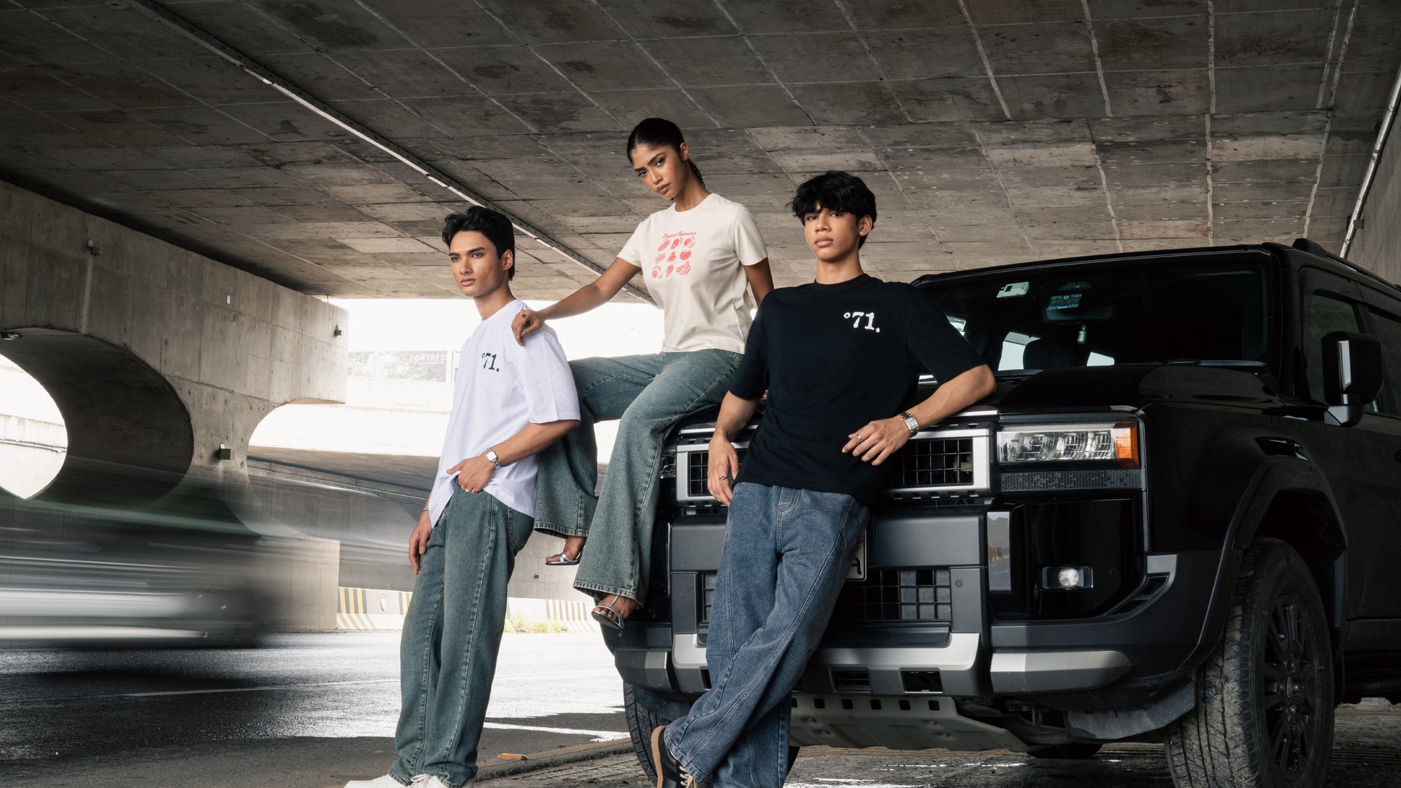 Three young men and women posing in Nº71 streetwear clothing in front of a black SUV under a concrete bridge.